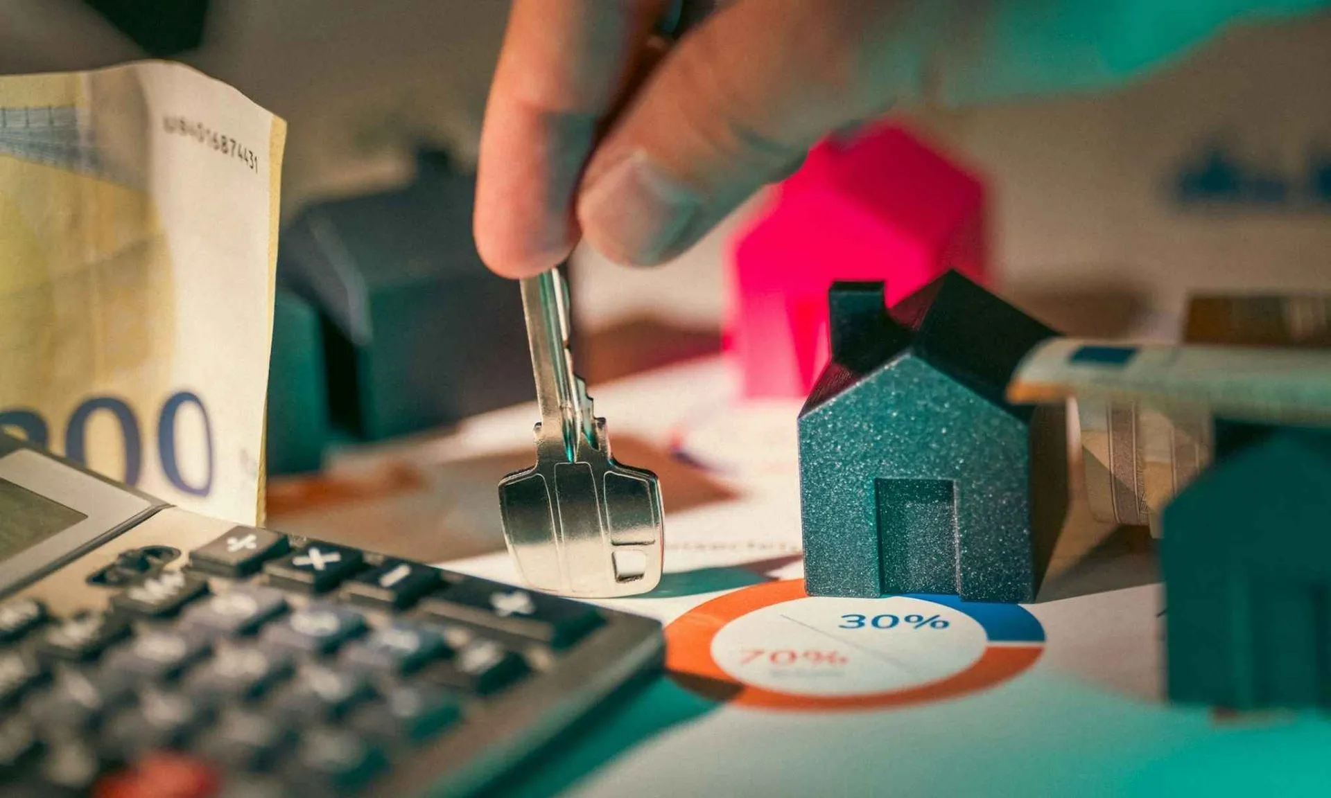 Hand holding key above miniature houses, with money, calculator, and pie chart.