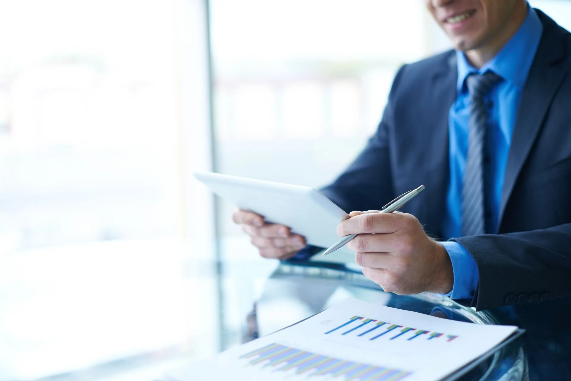 Man in suit reviews tablet and document with charts at a desk.