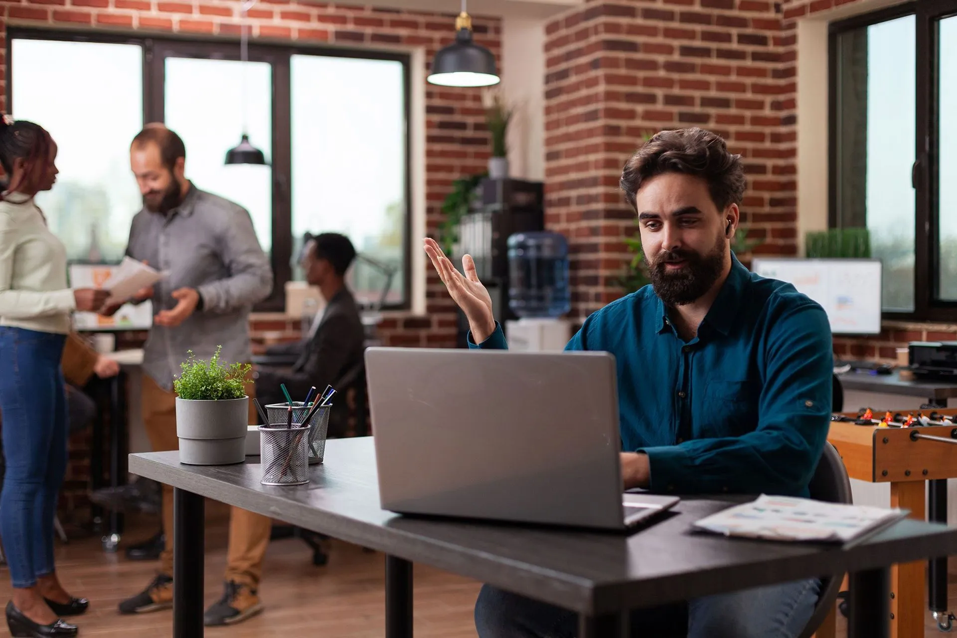 Man at laptop gestures while working in modern office. Others chat and work nearby.