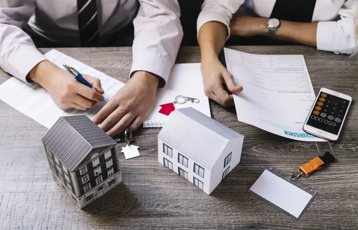 Two people at a table reviewing documents with house models, keys, and a calculator, discussing a real estate deal.