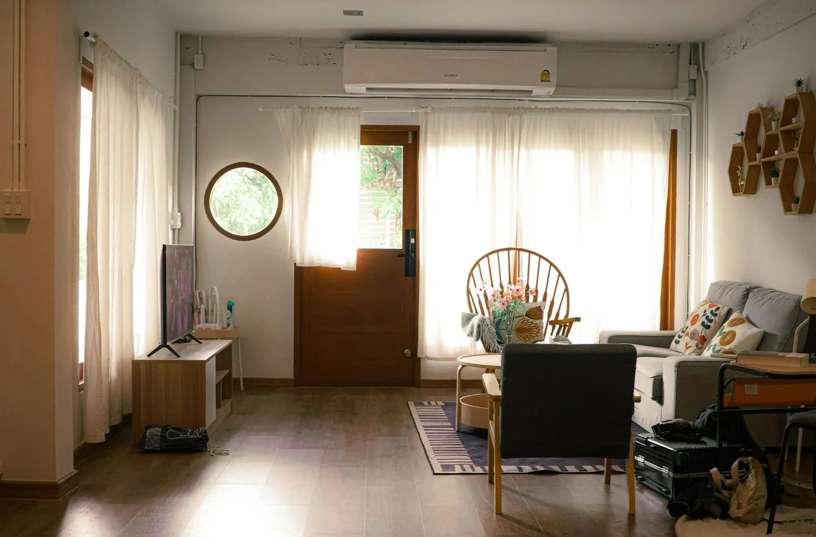 Cozy living room with wooden floors, neutral curtains, and natural light, including a sofa, chair, TV, and coffee table.
