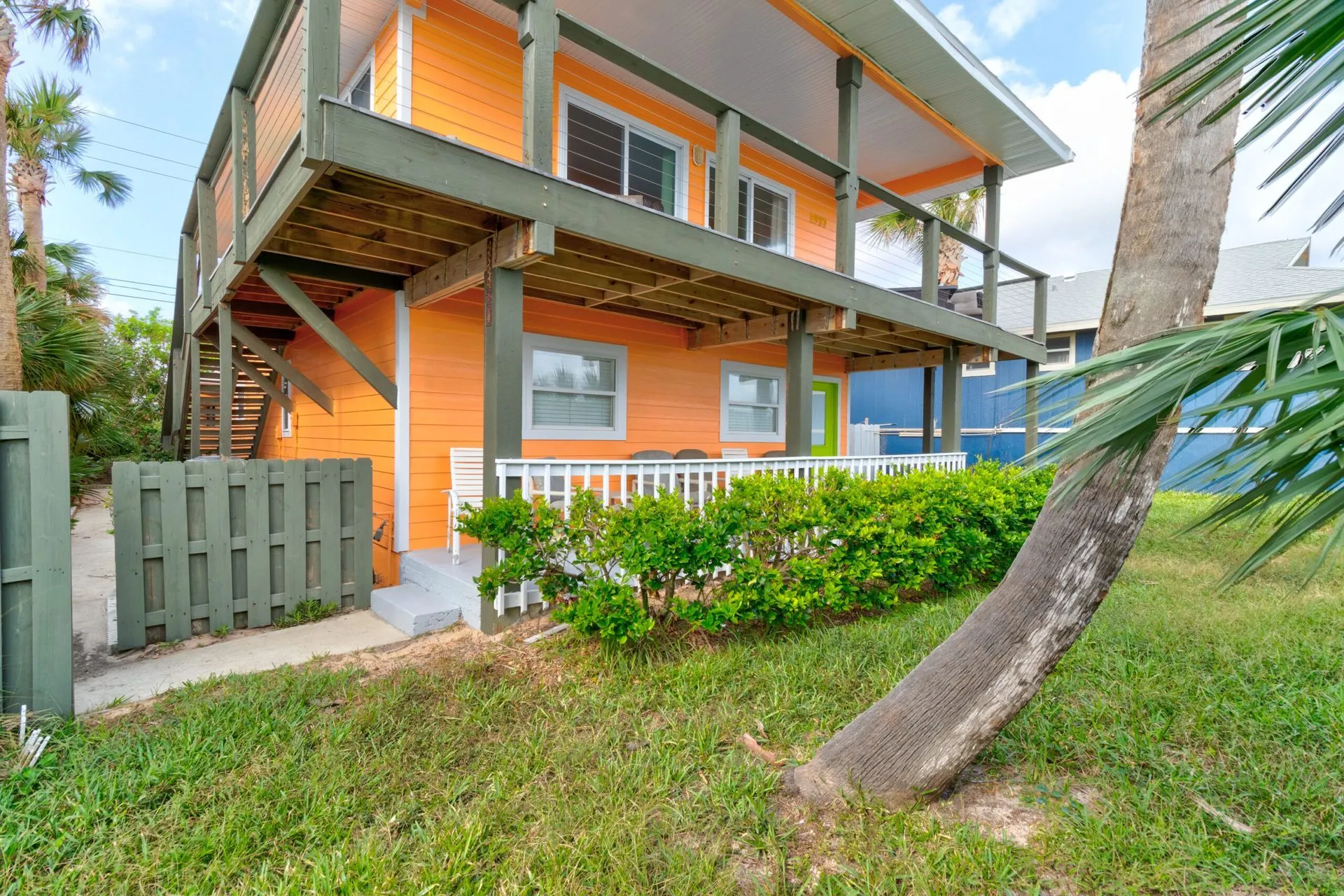 An orange beach house with a second-story deck, green trim, and white railing, near palm trees.