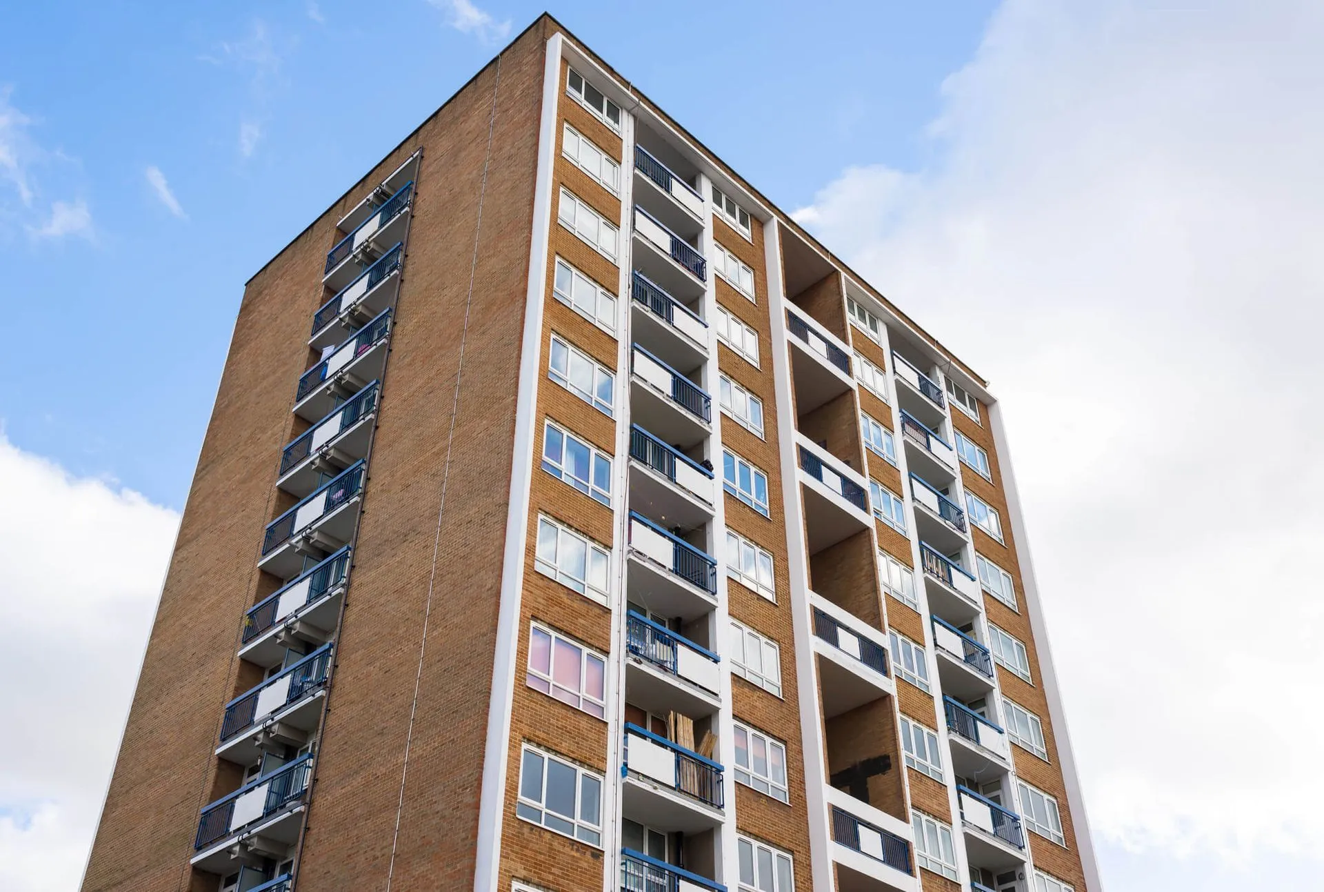 Tall brick apartment building with blue balcony railings against a cloudy sky.