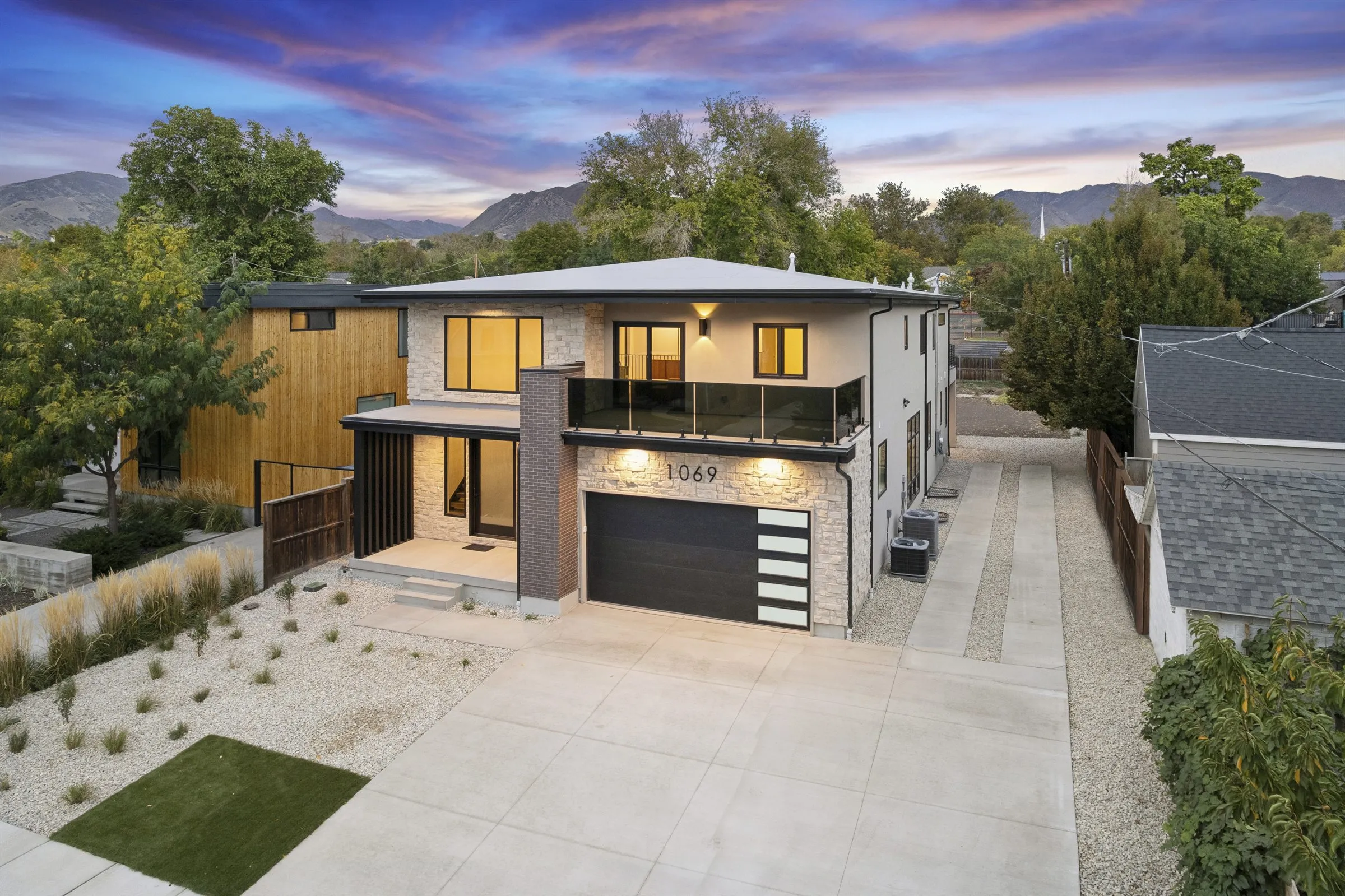 Aerial twilight view of a modern two-story home with a stone facade, glass balcony and two-car garage, with the Wasatch Mountains behind.