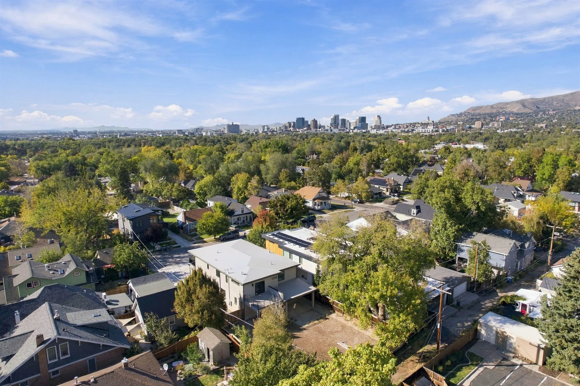 Daytime aerial view over a tree-lined Salt Lake City neighborhood of older bungalows and newer homes, with the downtown skyline and foothills behind.