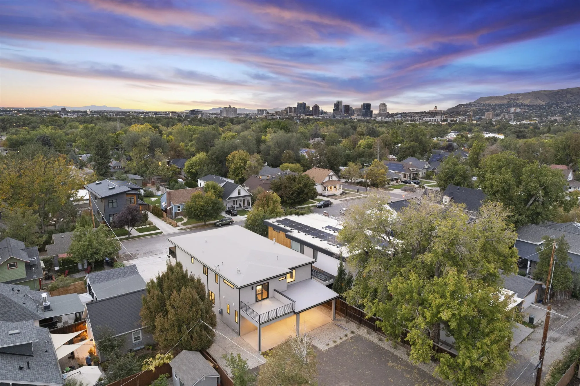 Twilight aerial view of a modern two-story home among older tree-lined Salt Lake City streets, with the downtown skyline and foothills at sunset.