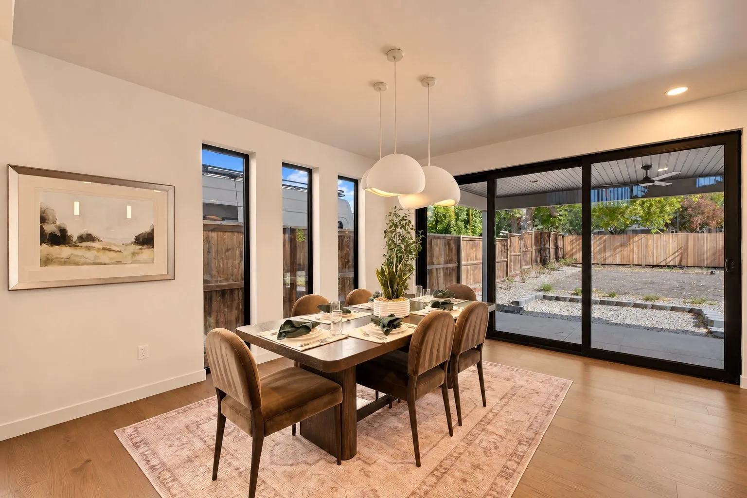 Dining room with a wood table, six velvet chairs, white pendant lights and sliding glass doors to a fenced backyard.